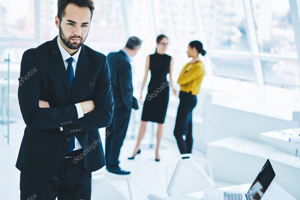 Confident man if elegant suit looking away while male and female colleagues on blurred background communicate about trade and accounting, serious bearded businessman thinking about idea for project