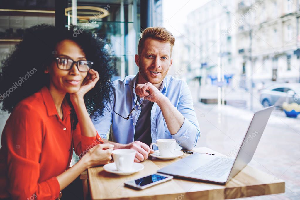 Portrait of positive multicultural male and female friends smiling at camera while spending leisure time togetherness for download movie on laptop computer with good internet connection in cafeteria