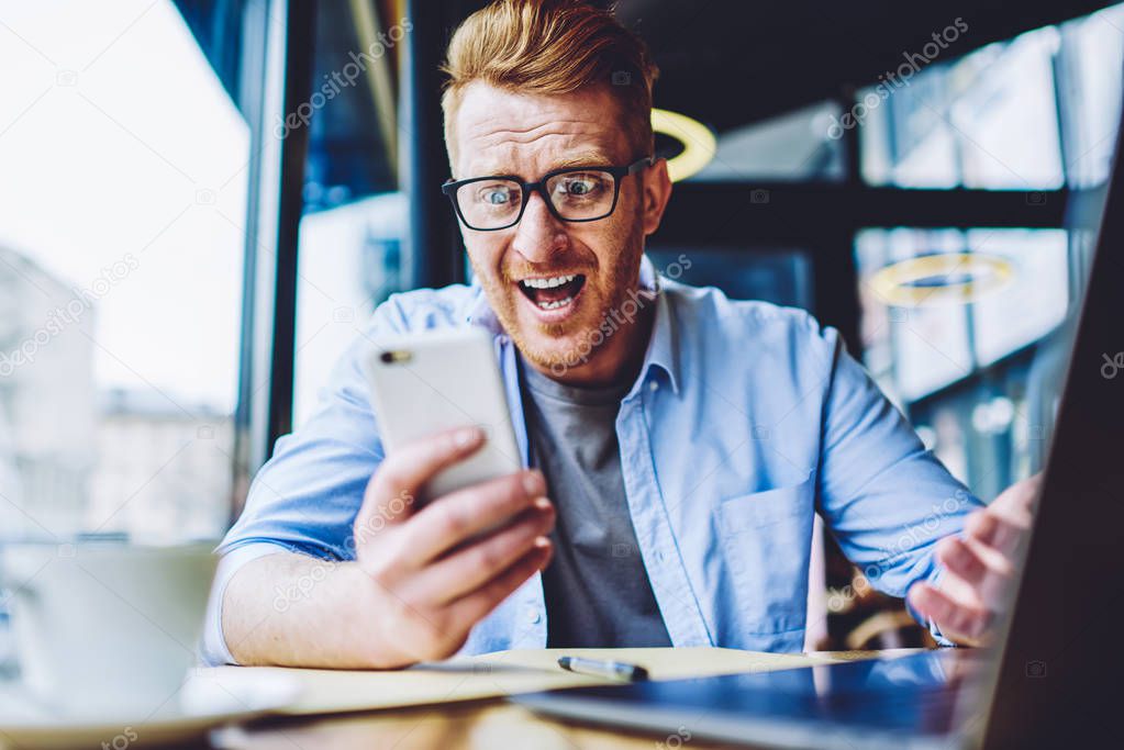 Amazed male blogger feeling shocked from price for application on cellular phone connected to public internet indoors, wonder hipster guy in spectacles checking notification on smartphone gadget