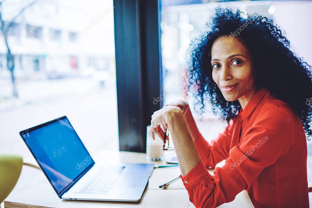 Portrait of cheerful female blogger sitting on publicity area with modern netbook and smiling at camera, positive woman dressed in casual apparel connected to 4g wireless on laptop computer