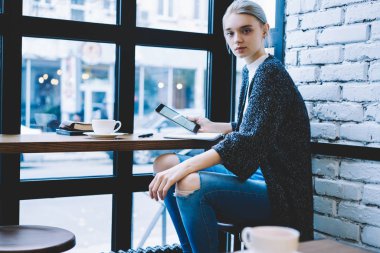 Oldukça modern sarışın saç kadın masada kahve dükkanında pencere yakın oturan ve dijital tablet tutarak. Millennial hipster kız harcama rekreasyon zaman kullanarak teknoloji 