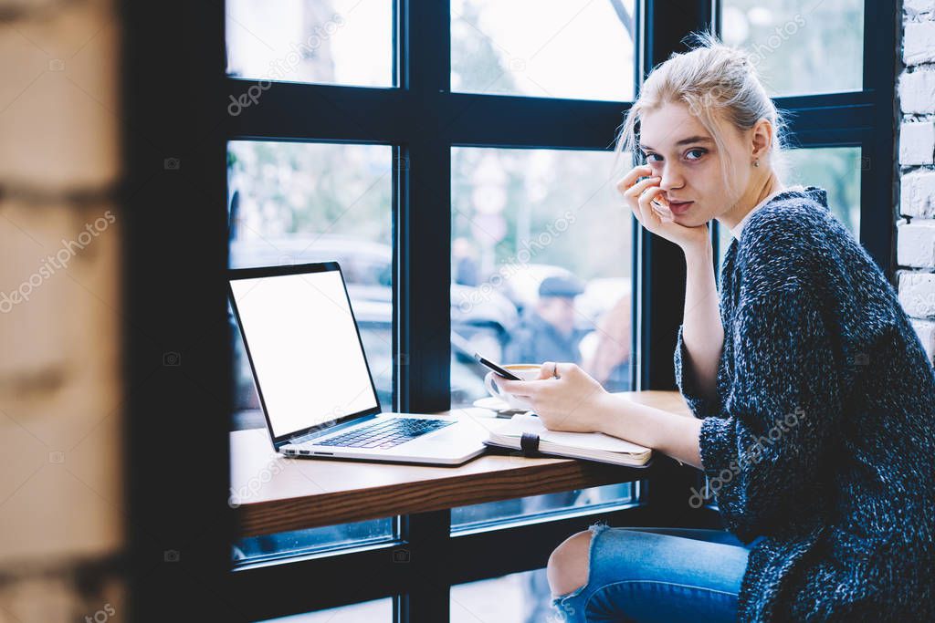 Young blonde hair female sitting with smartphone device in hands at the table with open laptop computer. Contemporary hipster girl spending time in coffee shop interior