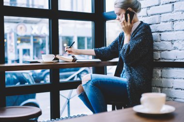 Kahve dükkanı iç masada otururken 20 yaşında kadın cep telefonu ile arama yan görünümü. Genç kadın Freelancer kullanarak teknoloji ve yazma notları Notepad