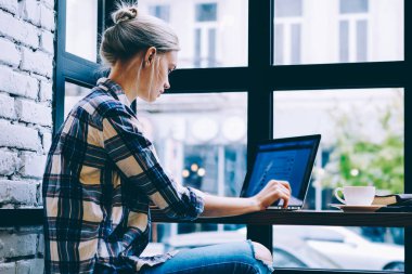 Pencere yakın yalnız oturan modern kahve dükkanı iç masada okurken dizüstü bilgisayar kullanarak Casual giysiler genç sarışın saç dişi yan görünümü. Freelance üzerinde çalışan genç çağdaş kadın