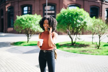 Karanlık tenli hipster öğrenci kahve molası sırasında akıllı telefon konuşma zevk açık havada, cep telefonunda uygulama üzerinden konuşurken rahat giyim kamera gülümseyen giyinmiş mutlu kadın