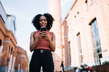 Kentsel ayarı üzerinde arkadaş beklerken bin yıllık hipster kız mobil uygulama üzerinden online oyunlar oynarken görünümü aşağıda, pozitif koyu tenli kadın cep üzerinden SMS mesajı göndermek için metin yazarak