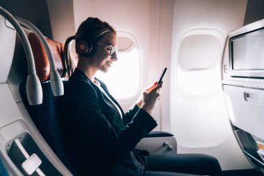 İş sınıfında oturan ve kulaklıklar olumlu ses kitabı dinlerken sohbet için gemide WiFi internet bağlantısı zevk popüler havayolları ile seyahat gülümseyen kadın yan görünümü