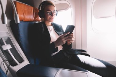 Resmi aşınma giyinmiş kafkas kadın yolcu koltuğuna oturan uçak içinde cep telefonu kullanımı, başarılı kadın okuma kulaklık ile ses kitabı dinlerken cep telefonu ile e-posta aldı