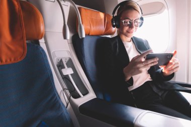 Pencere yakın oturan uçak uçuş sırasında akıllı telefonla okuma haberleri resmi aşınma Kafkas kız, Wi-Fi bağlantısı ve Bluetooth kulaklıklar kullanarak online video izlerken göz giyim iş kadını