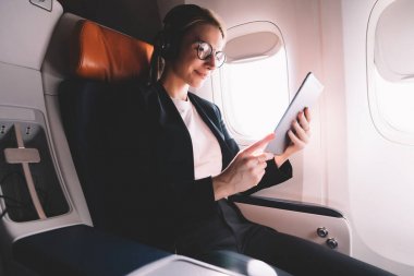 Pozitif genç kadın uçak yolcu koltuğunda oturan ve dijital tablet uygulama ve Bluetooth kulaklıklar üzerinden müzik şarkıları dinleme ile ilginç bir kitap okuma. Gemide kablosuz internet