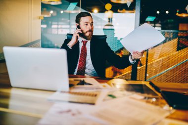 Evde ofis iç dizüstü bilgisayar ile masaüstünde oturan Smartphone çağrıda iken sakallı erkek gururlu CEO 'su elinde kağıt mali raporlar tutarak. Cep telefonunda konuşan ciddi işadamı