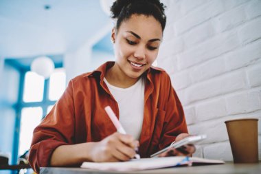 Pozitif Afro Amerikan kadın Blogger sosyal ağ sonraki yayınlar için ilginç ve komik fikirler için notlar yapmak, mutlu siyah öğrenci eğitim için mobil gadget ve ders kitabı ile okuyor