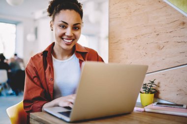 Kafeterya dizüstü cihaz ile boş zaman sırasında kamera gülümseyen mutlu kadın öğrenci portresi, yeni netbook ile masada oturan gündelik giyim müreffeh karanlık tenli hipster kız