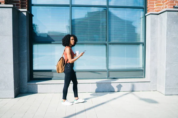 千禧一代时尚女孩穿着休闲装在城市街道上走来走去 手里拿着外卖杯和智能手机小工具 黑皮肤的女学生用现代科技在城市环境中漫步 — 图库照片