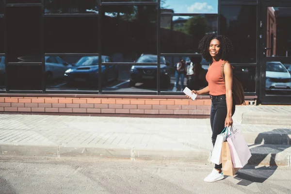 城市阳光明媚的日子 快乐的女性购物狂在城市环境中漫步 手握好购物品 面带微笑 非洲裔美国买家带着纸袋 购物时感觉良好 — 图库照片