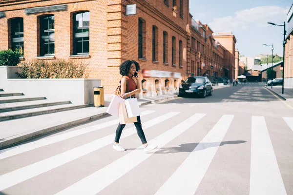 快乐的非洲裔美国女性买家与购物袋在城市设置过马路 并打电话给朋友夸耀伟大的购买 积极的女人在休闲装交谈通过应用程序 — 图库照片