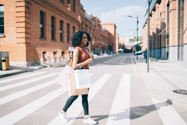 Smiling female teenager trendy dressed crossing road and communicating with customer service for discussing good sales of Black Friday, happy woman enjoying shopping time in city using smartphone