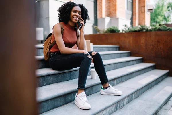 Portrait of cheerful female student dressed in casual apparel smiling at camera while communicating via mobile application, positive millennial black model using roaming connection on cellular