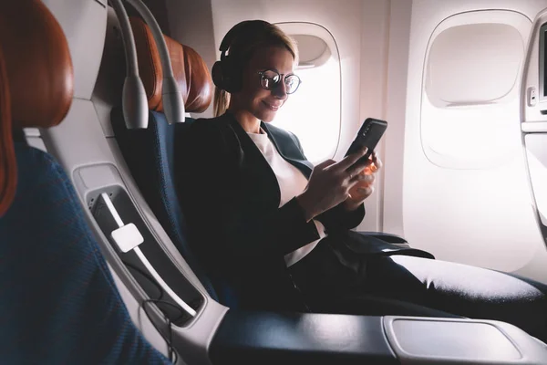 Smiling female passenger using smartphone connecting to wireless internet in airplane cabin, caucasian young woman traveler enjoying wifi hotspot service on board during travel time