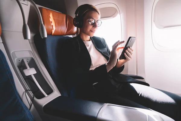 年轻的白人妇女坐在靠近窗口与智能手机连接到Wifi互联网在飞行期间在机上 女乘客在耳机的噪音取消搜索歌曲在在线商店的应用程序 — 图库照片