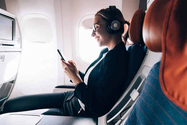 Cheerful female passenger in headphones for noise cancellation watching online movie during intercontinental flight in cabin of aircraft,happy young woman using wifi connection on board, playing games