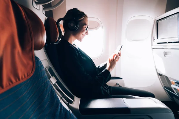 飛行中に携帯電話で音楽を聴いたり ネットワークを閲覧したりして 飛行機の助手席に座っている陽気な女性音楽愛好家の側面図 機内でワイヤレスを使用する若い女性の乗客 — ストック写真