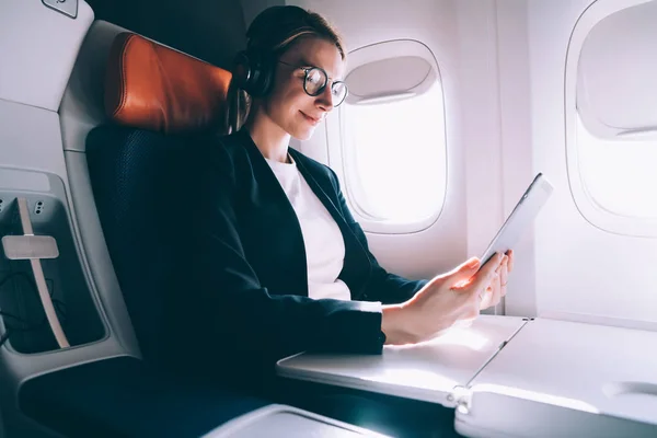 迷人的女性在蓝牙耳机噪音消除观看在线网络研讨会在舒适的飞行在飞机上 妇女浏览网络的数字平板电脑 而连接到无线热点 — 图库照片