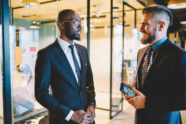 Two successful multicultural office employees in suits discussing talking while standing in office interior. Prosperous diverse entrepreneurs having friendly conversation about work