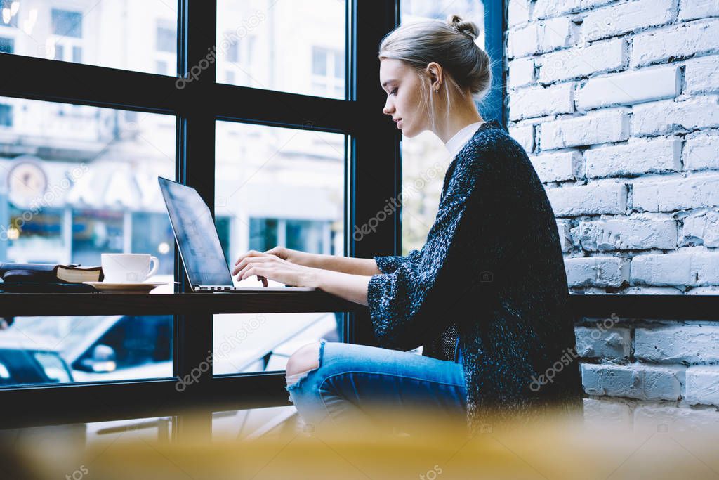 Side view of young blonde hair female in casual clothes using laptop while studying at table in modern coffee shop interior sitting alone near window. Young contemporary woman working on freelance