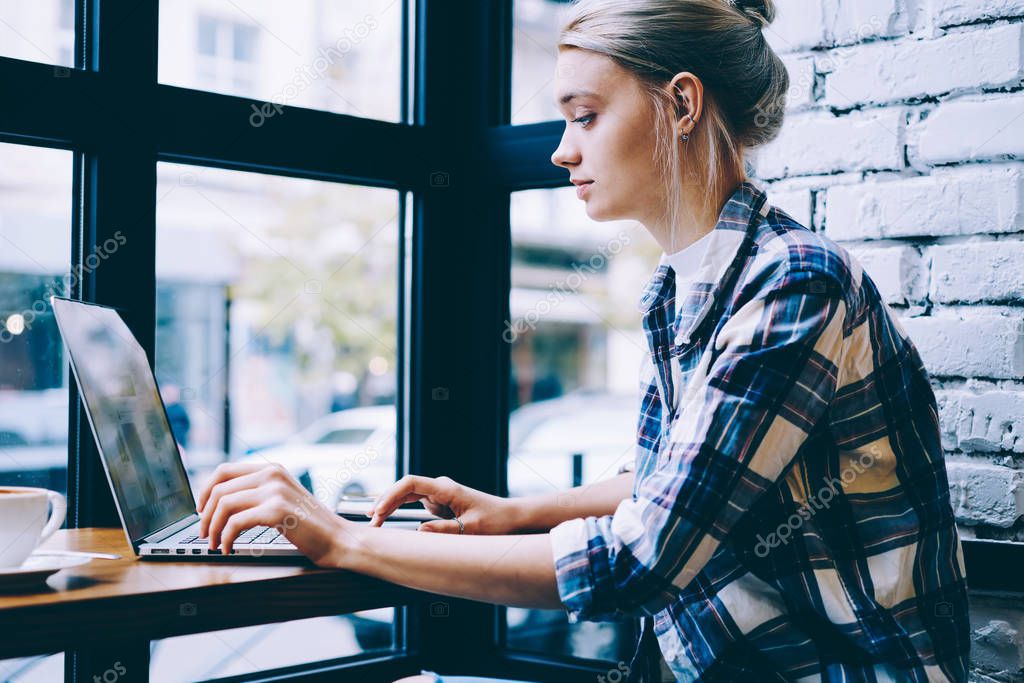 Side view of young blonde hair female in casual clothes using laptop while studying at table in modern coffee shop interior sitting alone near window. Young contemporary woman working on freelance