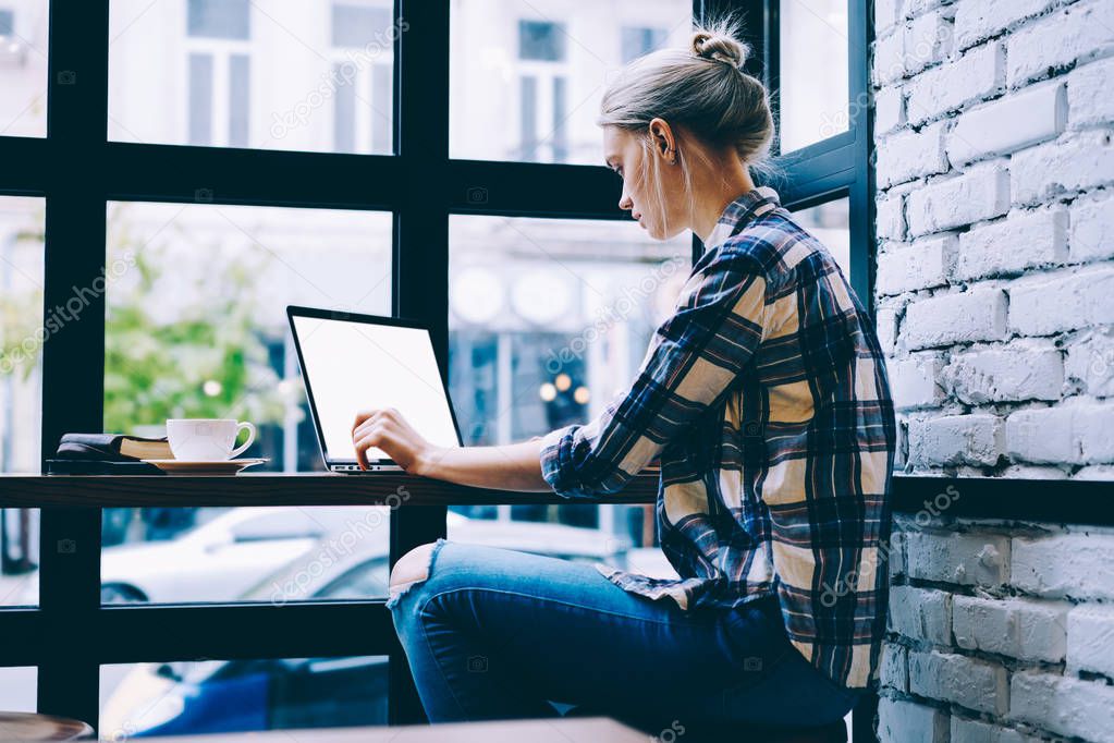 Side view of young blonde hair female in casual clothes using mock up laptop while studying at table in modern coffee shop interior sitting near window. Young contemporary woman working on freelance