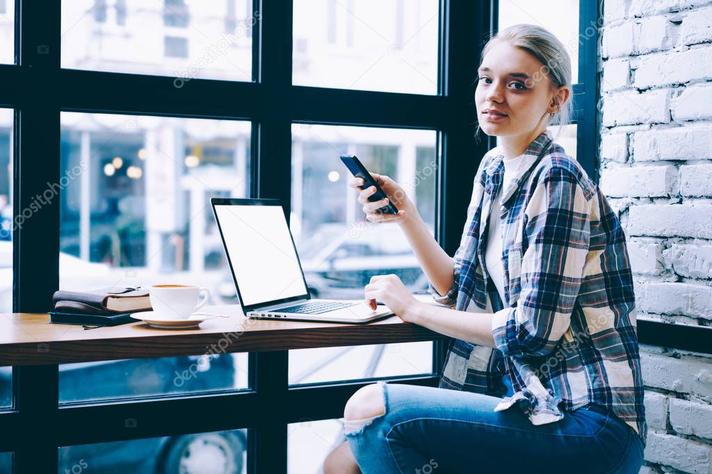 Contemporary blonde hair female sitting at table near mock up laptop computer browsing network via application on mobile phone. Millennial hipster girl spending time using technology in coffee shop