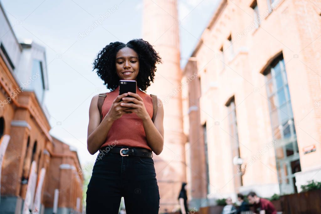 Below view of millennial hipster girl playing online games via cellular application while waiting friend on urban setting, positive dark skinned woman typing text for sending sms message via mobile