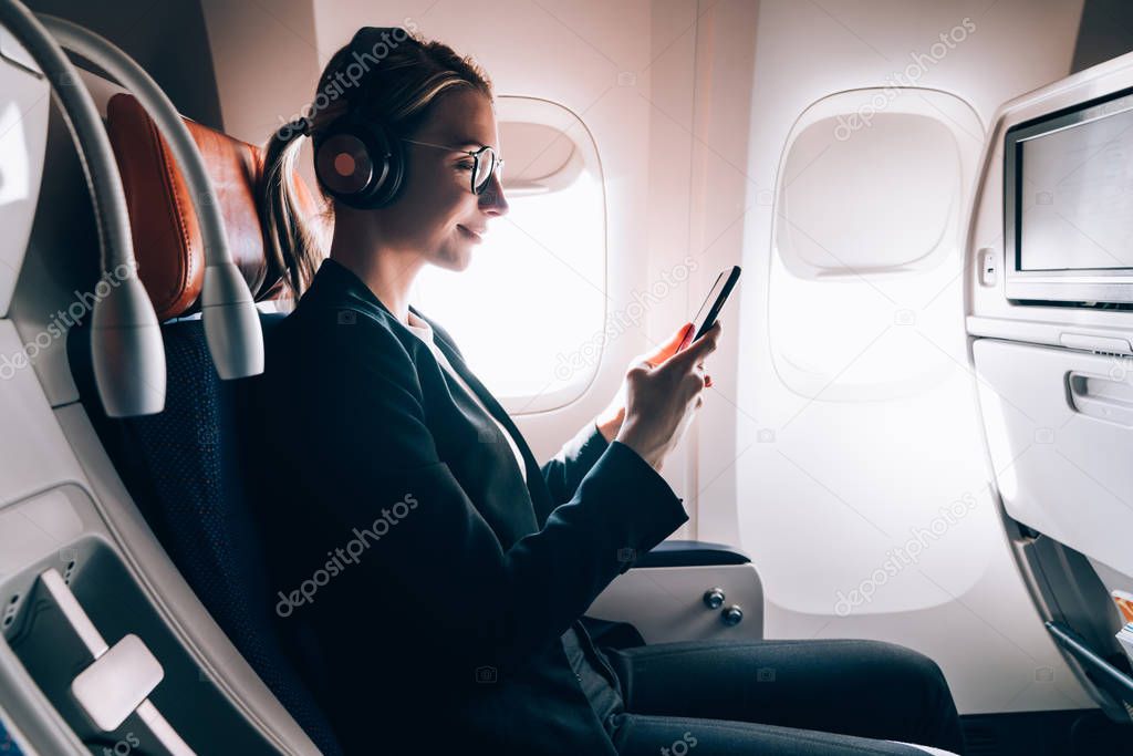 Smiling female passenger in bluetooth headphones for noise cancellation chatting online via mobile phone using wifi internet connection on board. Young woman in seat near window using smartphone app