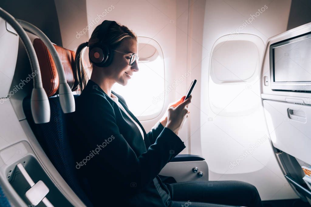 Side view of smiling female travelling via popular airlines sitting in business class and enjoying wifi internet connection on board for chatting while listening positive audio book in earphones