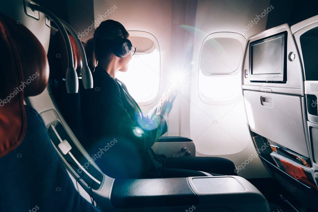 Side view of businesswoman in elegant wear looking in airplane window and waiting take-off from airport during trip to conference, intelligent female in electronic headphones listening audio book