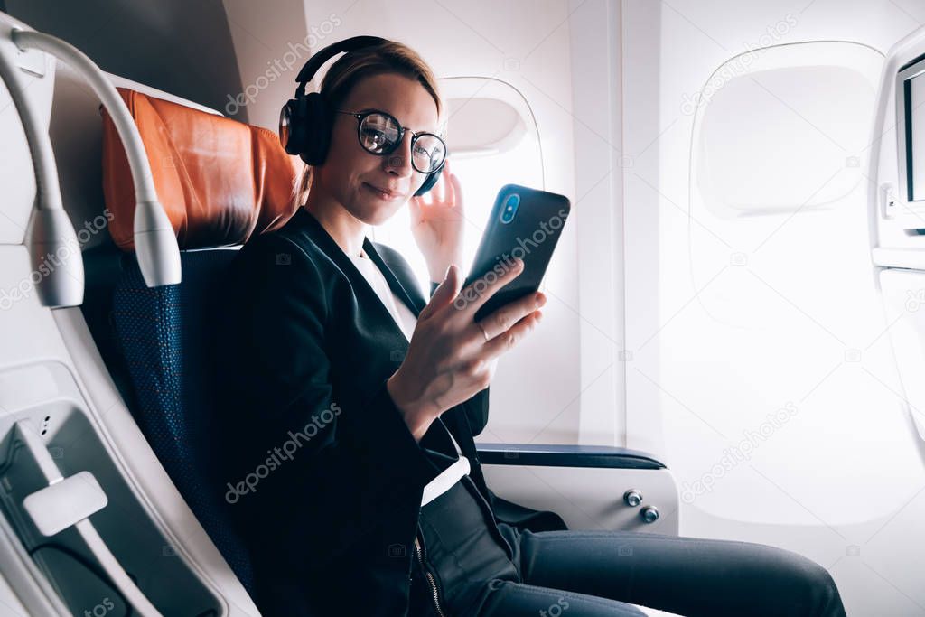 Travel tourism technology and air flights concept, intelligent woman sitting in plane with modern smartphone gadget and searching favourite music playlist in application for melomans for listening