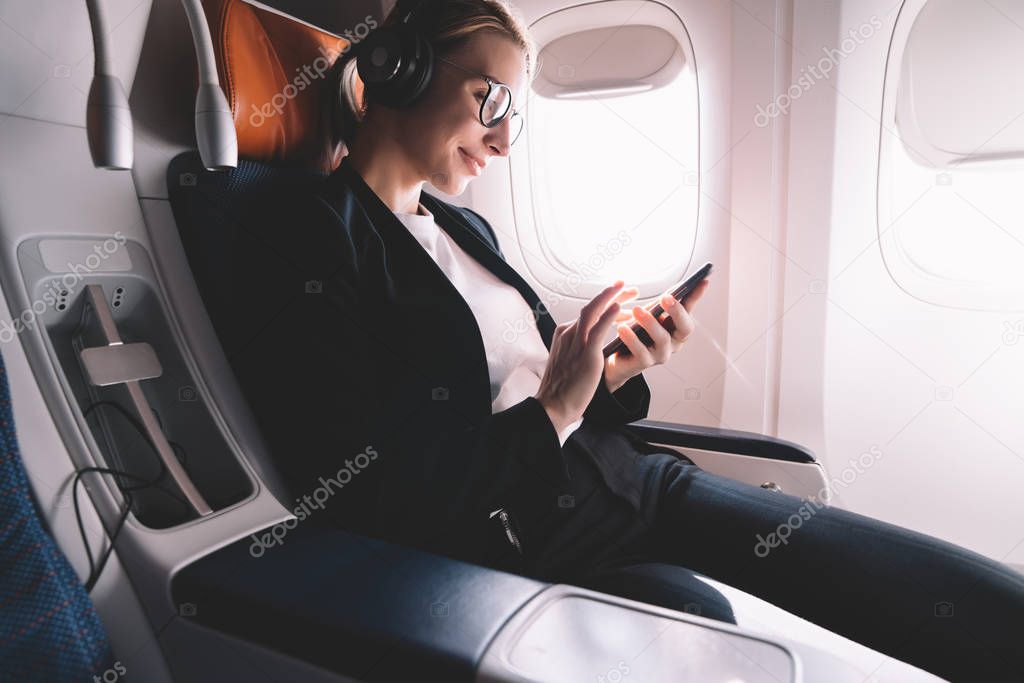 Hipster girl flight passenger connected by smartphone to wireless network inside airplane cabin, smiling female in bluetooth headphones for noise cancellation enjoying music songs on mobile phone app