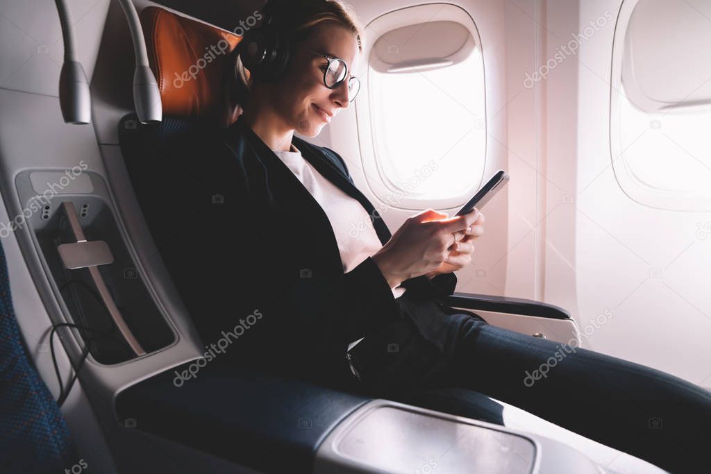 Happy female passenger using mobile phone searching WiFi Connection on board in airplane, successful lawyer in formal suit chatting online via application on cellular while enjoying music