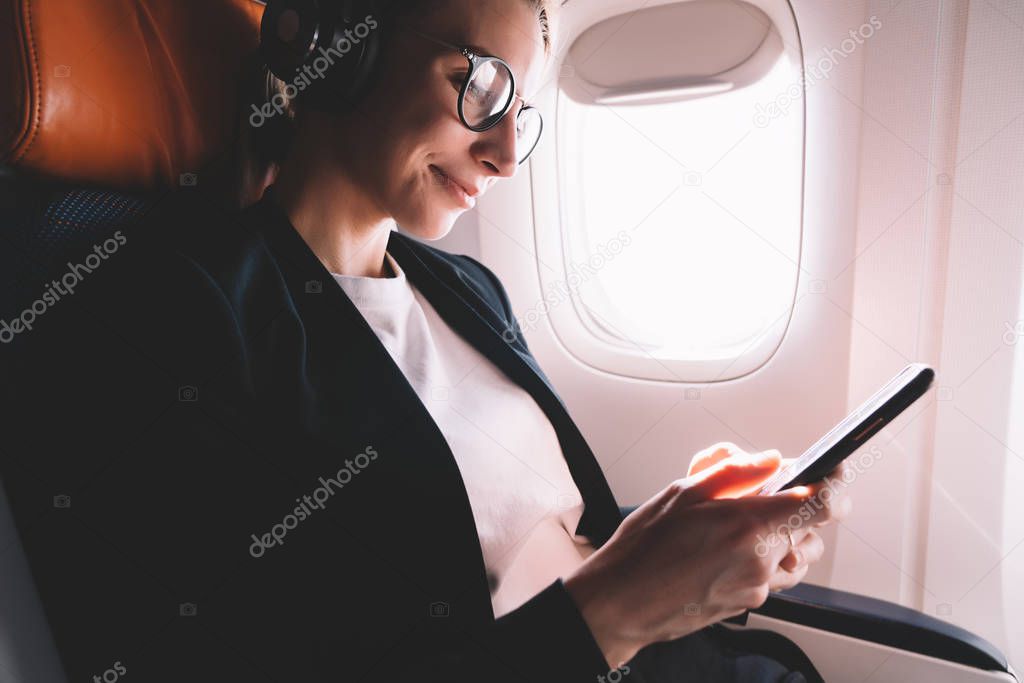 Happy smiling hipster girl listening music with cellphone on airplane connected to wifi on board, positive female passenger enjoying flight in first class with good wireless internet on smartphone 