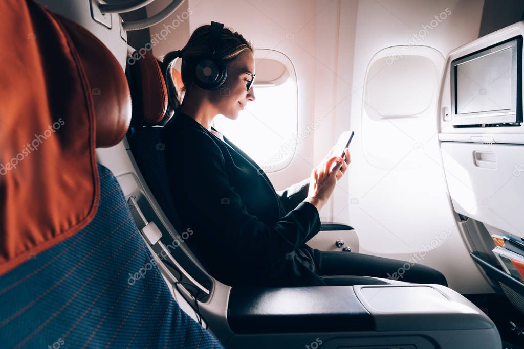 Side view of woman in formal wear watching online positive video while using wireless internet connection on board, female passenger reading news while listening audio record via headphones