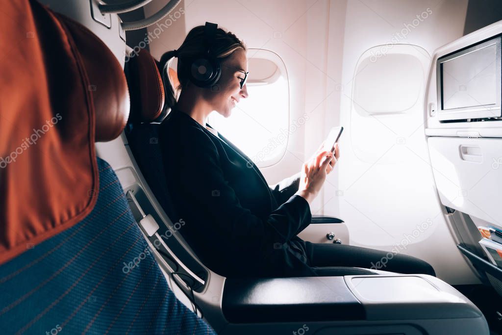 Side view of cheerful female music lover sitting in passenger seat in airplane enjoying listening music and browsing network via cellphone during flight.Young woman passenger using wireless on board