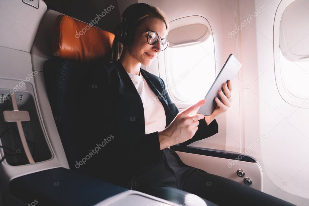 Positive young woman sitting in passenger seat in airplane and reading interesting book via application on digital tablet and listening music songs via bluetooth headphones. Wireless internet on board