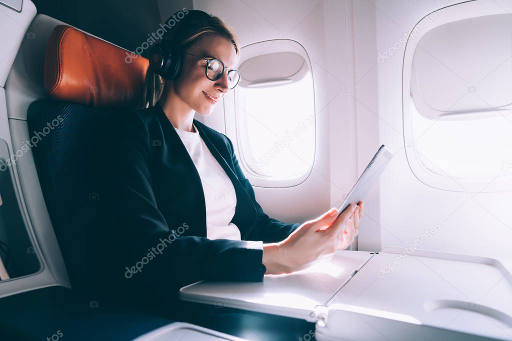 Positive woman in spectacles feeling good from online video call to friend during intercontinental flight in first class with good wifi connection on board, concept of aircraft and airline
