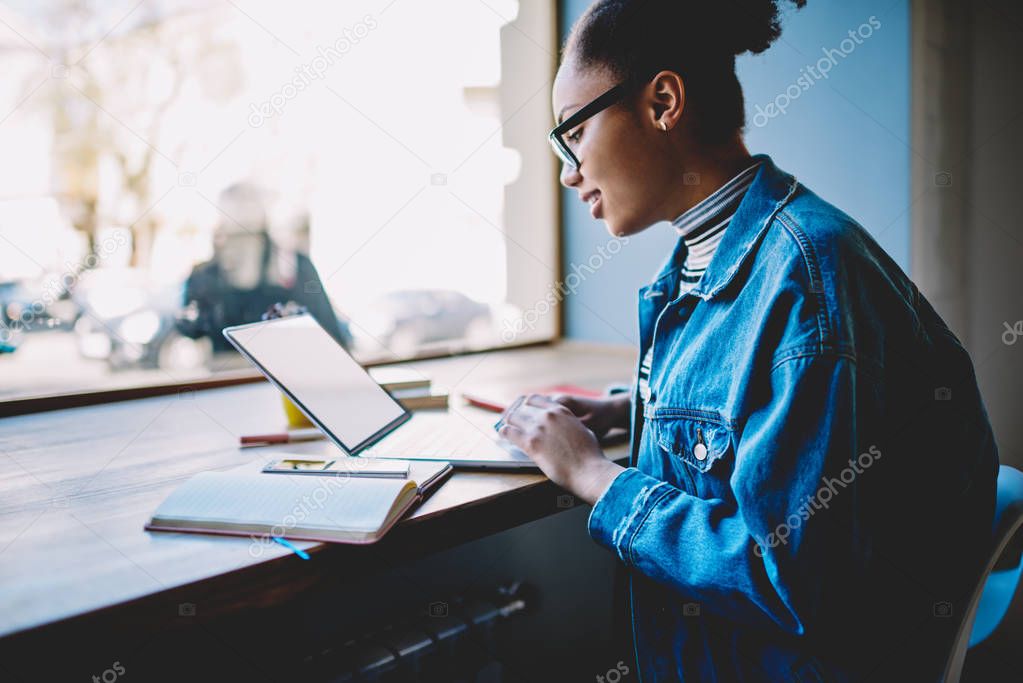 Side view of positive female student using public internet for making shopping on web store during break from exam preparation, dark skinned skilled woman searching information for course work