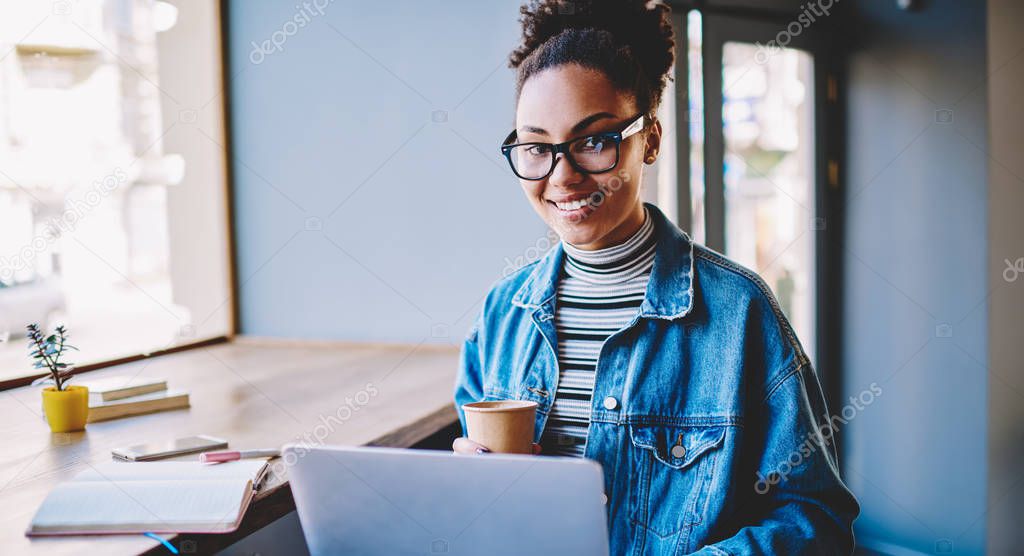Portrait of happy black female student sitting in university cafeteria with takeaway cup and enjoying coffee break from e learning and research of data on laptop computer, concept of autodidact