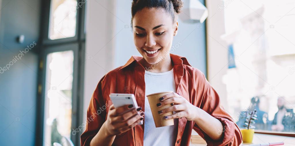 Happy dark skinned hipster girl sending sms message via smartphone device while enjoying caffeine beverage, african woman holding paper cup with coffee and reading article on content funny website