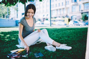 Tasarım okulunda muayene için hazırlanırken pozitif kadın mimar cep telefonu zaman kontrol, güneşli parkta mutlu gülümseyen hipster kız harcama zaman ve açık havada öğrenme zevk