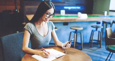 Gözlük pozitif kadın öğrenci cep telefonu ücretsiz kablosuz bağlantı kullanarak üniversite ödevleri yapmak için bilgi arama, mutlu hipster kız sosyal ağda olumlu haber okuma