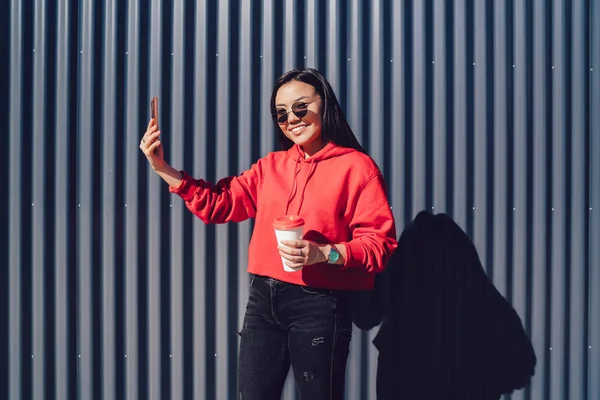Half length portrait of cheerful chinese female blogger in sunglasses posing near advertising area with coffee to go in hand and smiling at camera while clicking selfie photos on modern cellphone